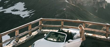 A white convertible car parked on a road with a wooden railing, overlooking a mountainous landscape with snow patches.