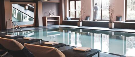 A modern indoor swimming pool with lounge chairs and a view of the outdoors.