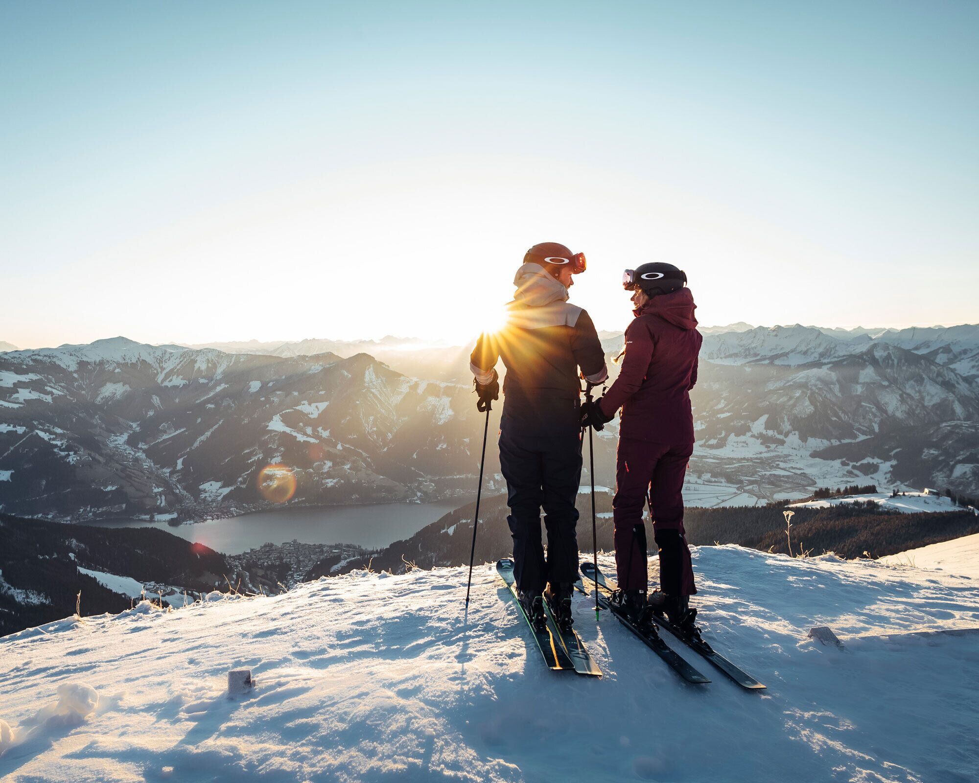 Two people skiing on a snowy mountain with a lake and mountains in the background.