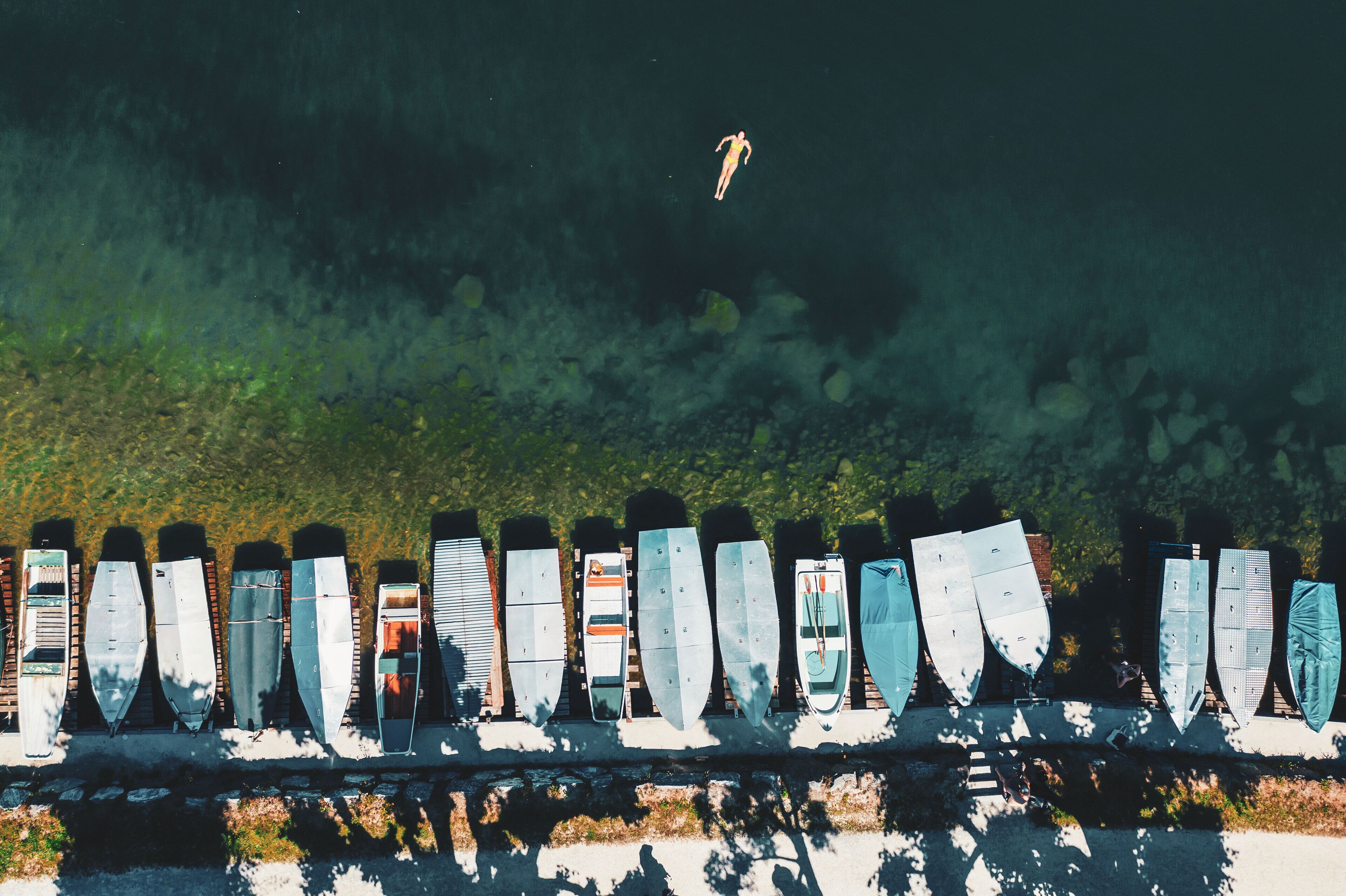 Eine Reihe von Booten, an einer Mauer entlang gereiht, mit einer Person, die dahinter im Wasser schwimmt.