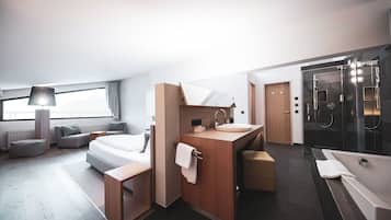 Una habitación de hotel moderna con una cama grande, una zona de estar con sofá y sillón, un tocador con espejo y lavabo, y un área de ducha separada.