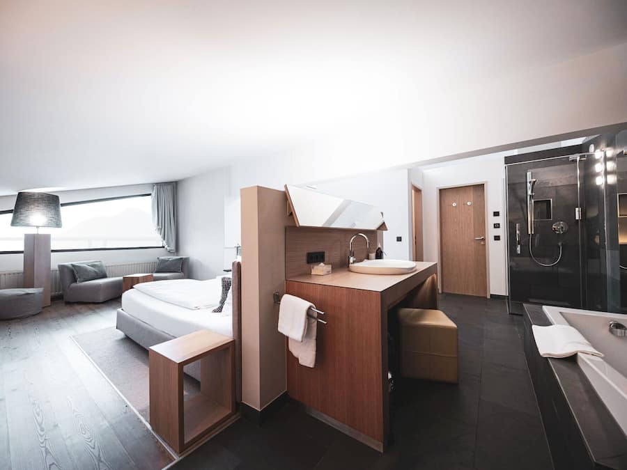 Una habitación de hotel moderna con una cama grande, una zona de estar con sofá y sillón, un tocador con espejo y lavabo, y un área de ducha separada.