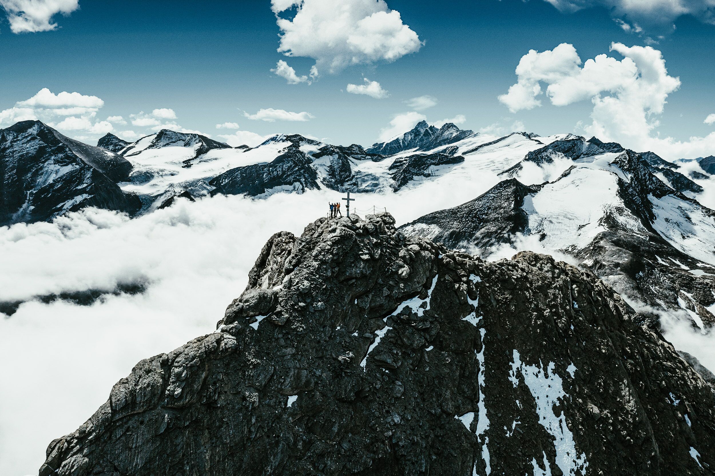Un pico montañoso con una cruz en la cima, rodeado de picos nevados y nubes.