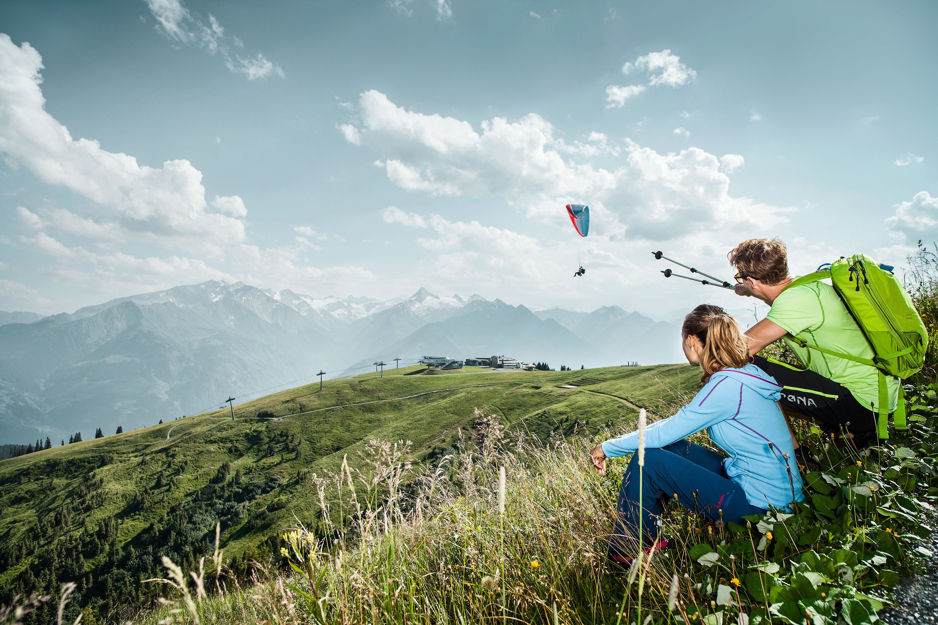 A person with a green backpack sitting on a grassy hill overlooking a mountainous landscape with a kite in the sky.