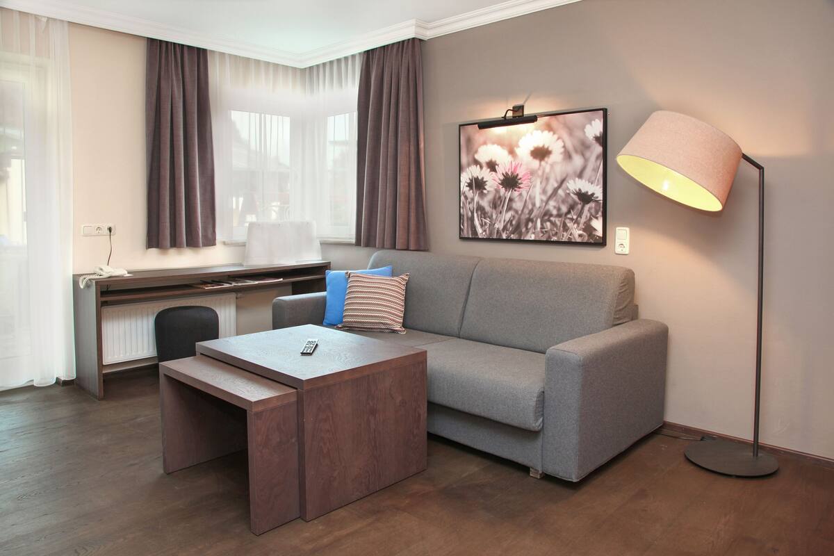 Ett modernt hotellrum med en grå soffa, ett träbord, ett skrivbord med en stol, en golvlampa och ett inramat konstverk med blommotiv på väggen.