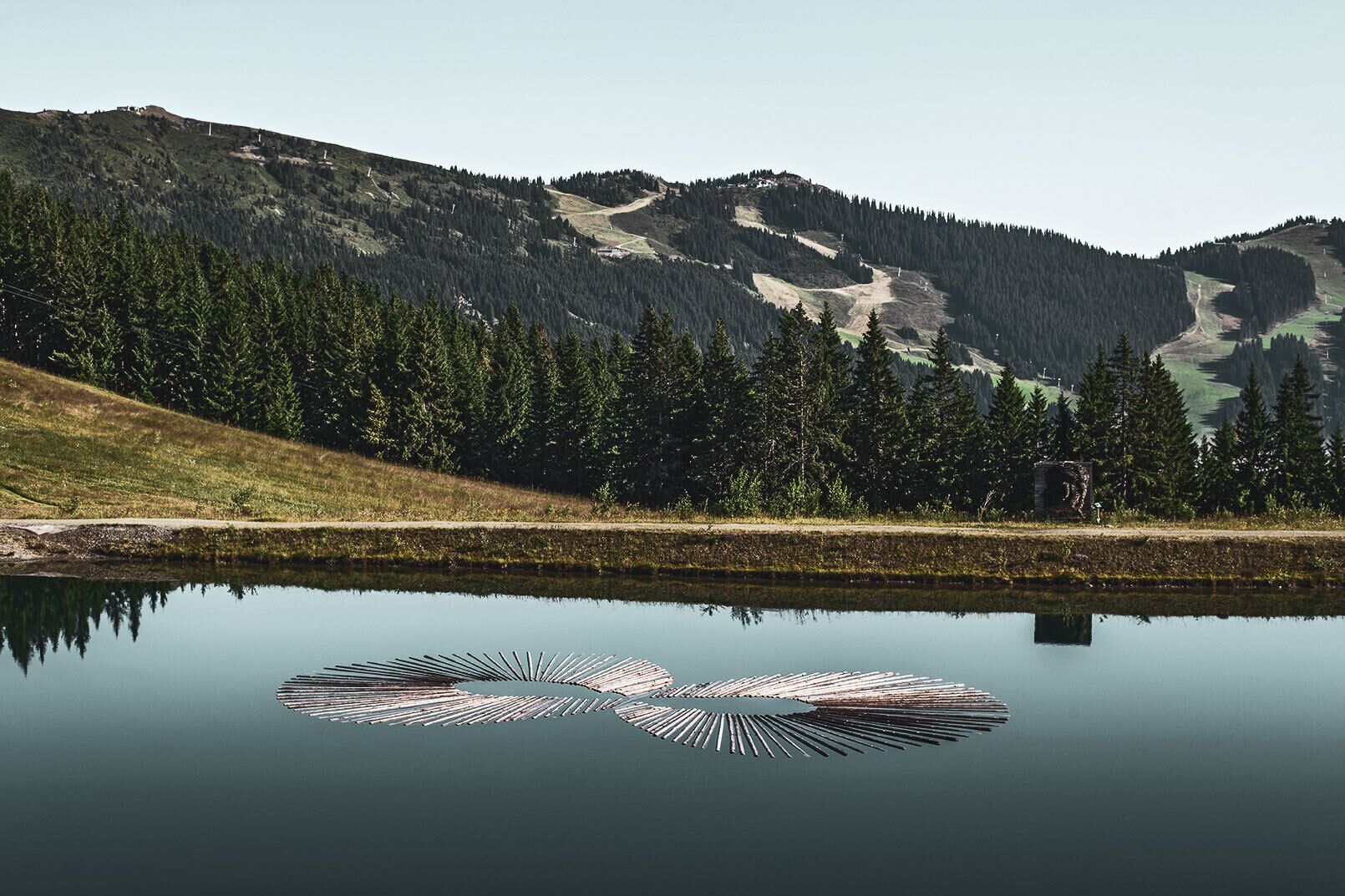 A calm lake with a symmetrical pattern of logs in the center, surrounded by a forested mountain landscape.