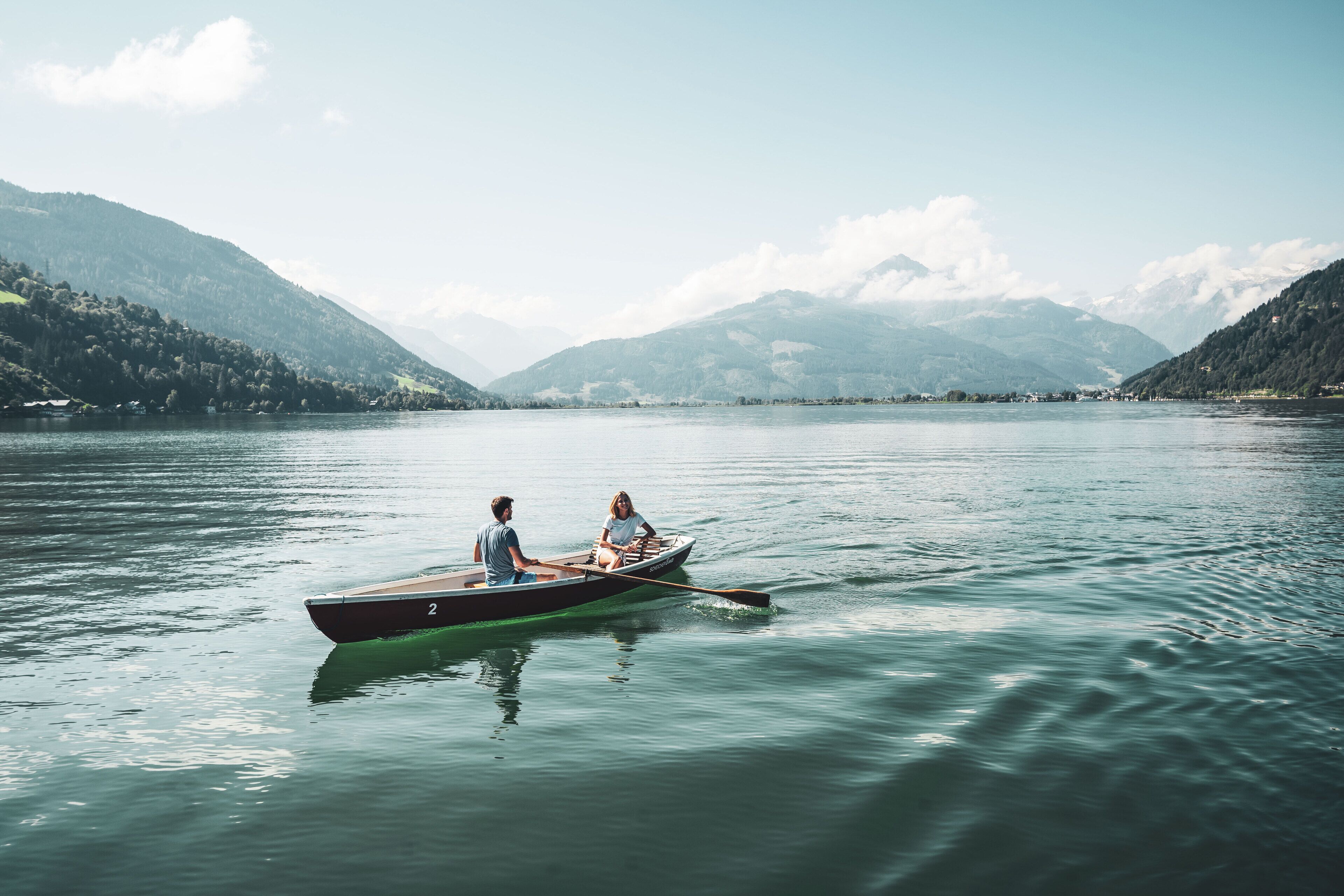 A calm lake with a rowboat carrying two people, surrounded by mountains and clear skies.