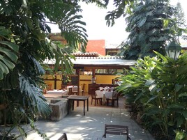 Un patio al aire libre con mesas y sillas, rodeado de exuberante follaje y un edificio con tejado de tejas.