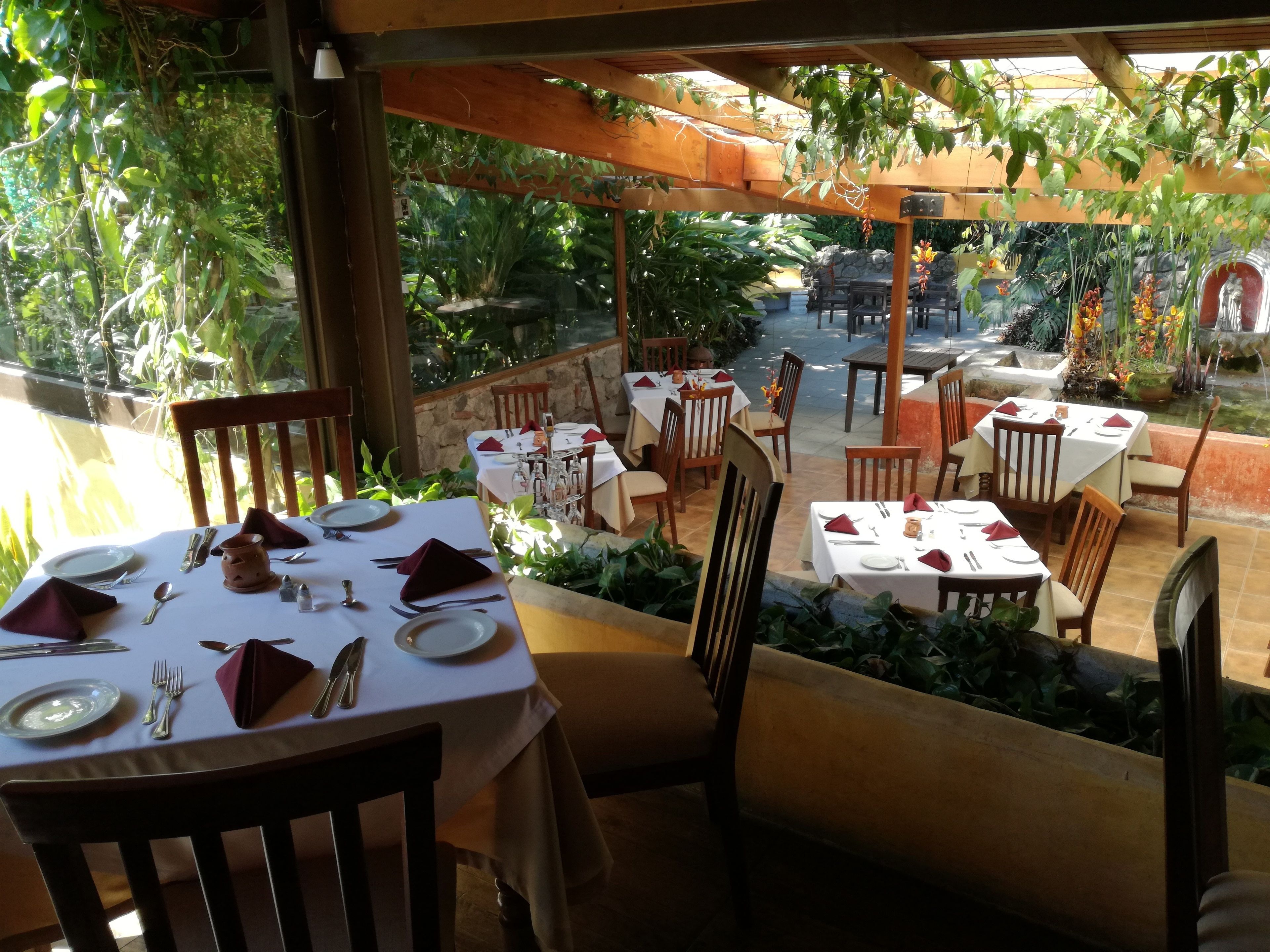 An outdoor dining area with tables set for two, surrounded by greenery and a stone wall.