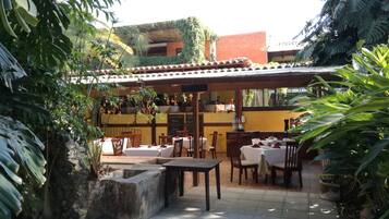 Un patio al aire libre con mesas y sillas, rodeado de plantas tropicales y una pérgola.
