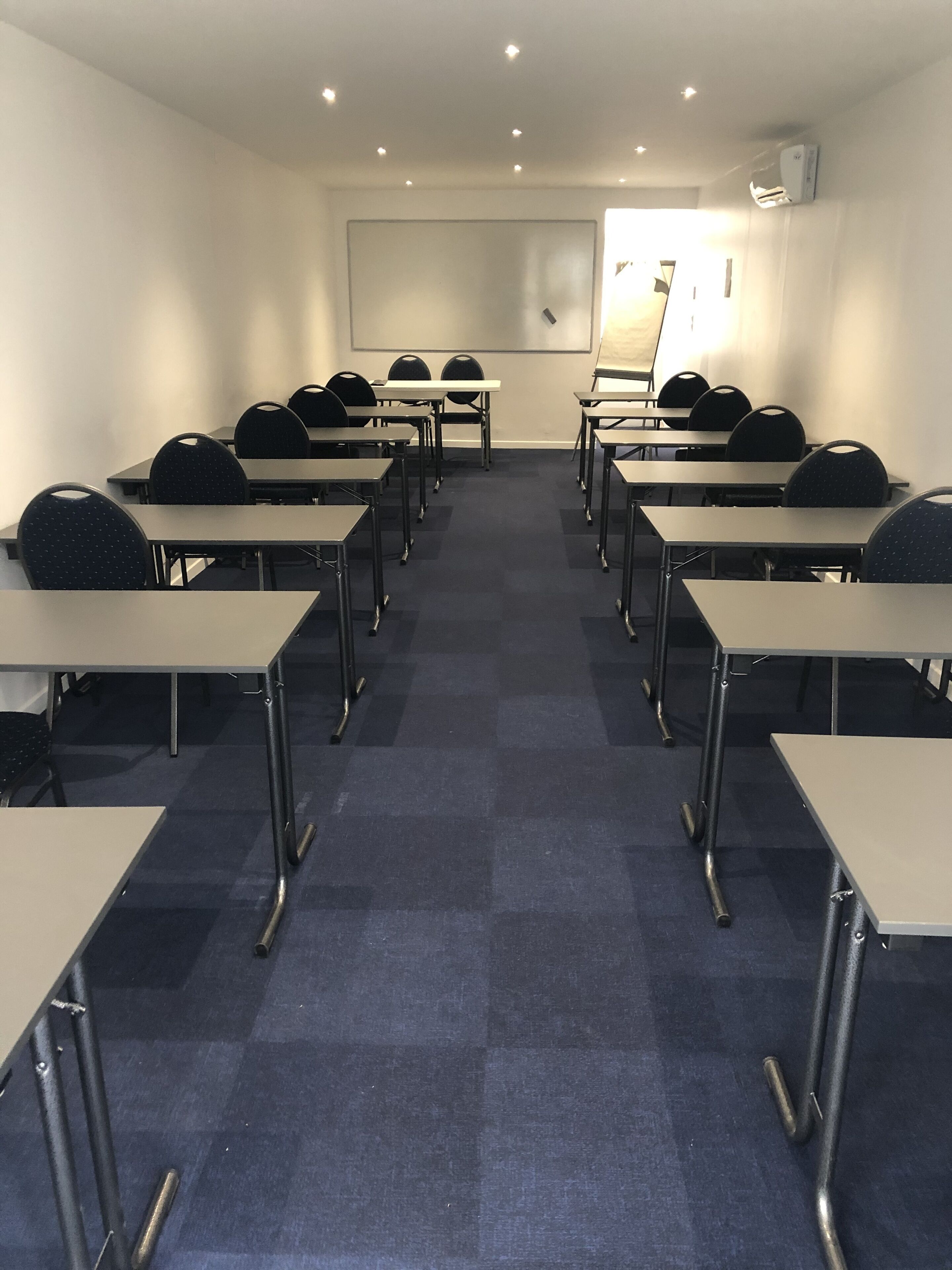 Een congreszaal met rijen tafels en stoelen, een projectiescherm en een airconditioner aan de muur.
