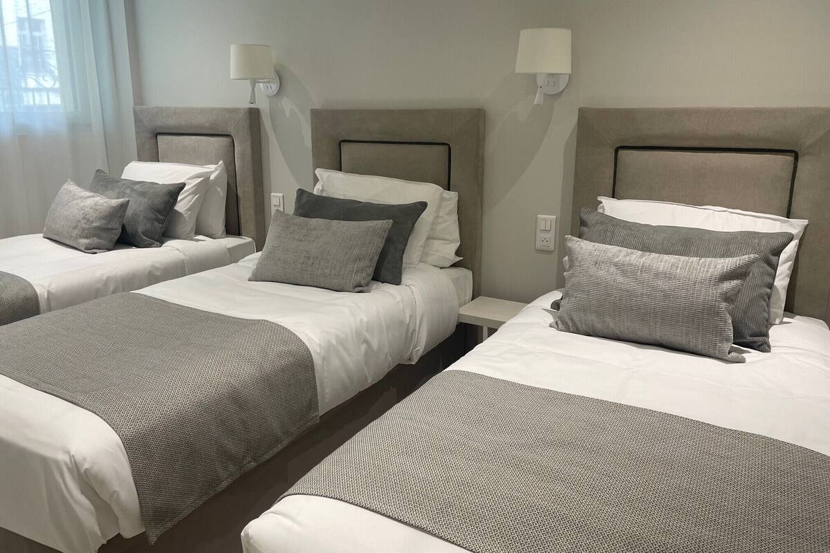 A hotel room with three beds, each with a headboard, bedside lamps, and grey bedding.