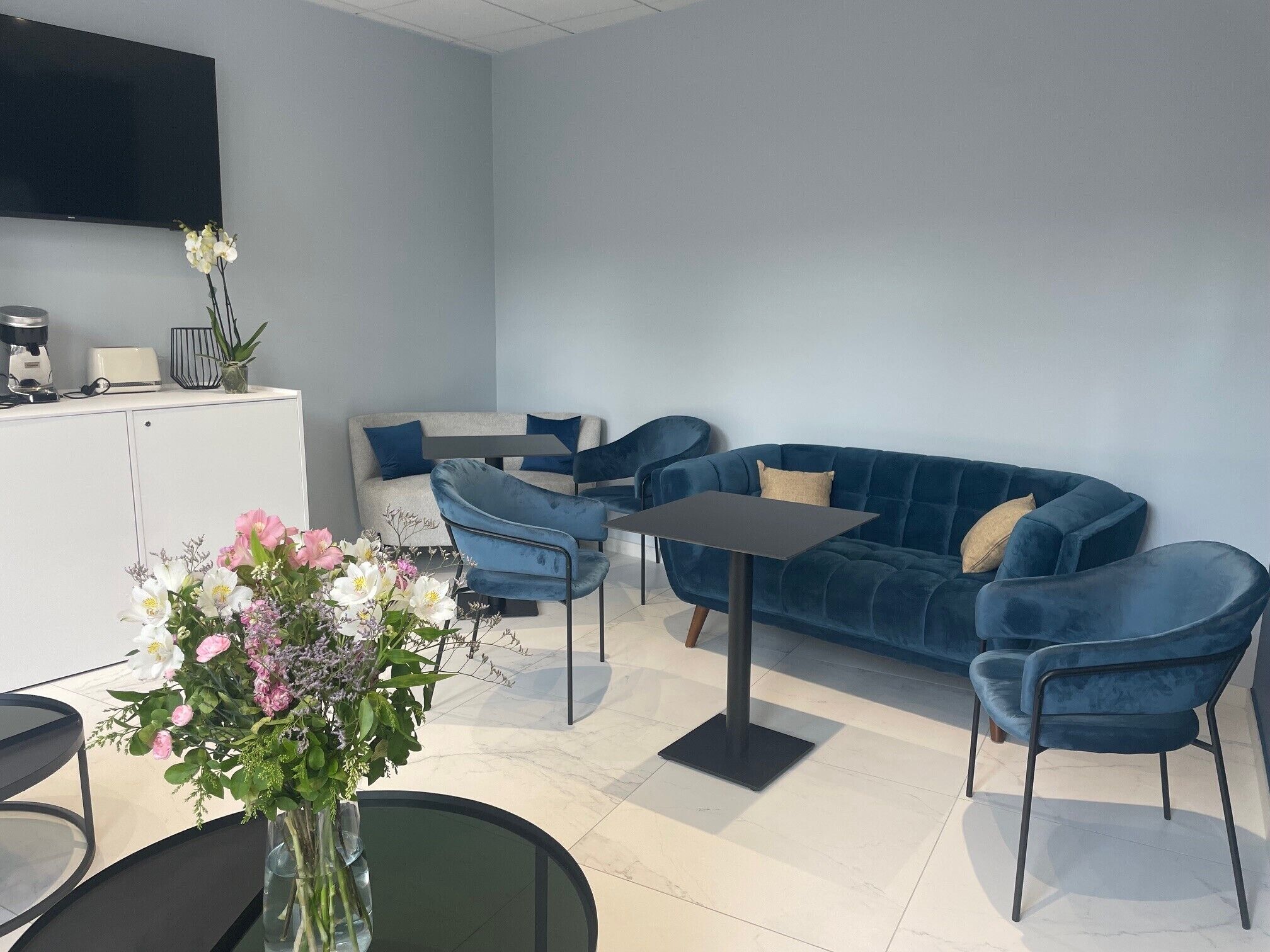 Un salon moderne avec un canapé bleu, deux fauteuils bleus, une table basse et une table à manger. Un téléviseur à écran plat est fixé au mur, un vase avec des fleurs est posé sur une table d’appoint, et un coin cuisine comprend un grille-pain et une machine à café.
