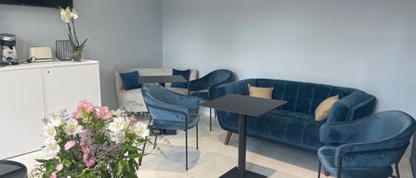 A modern living room with a blue sofa, two blue armchairs, a coffee table, and a dining table. There is a flat-screen TV mounted on the wall, a vase with flowers on a side table, and a kitchenette area with a toaster and coffee maker.