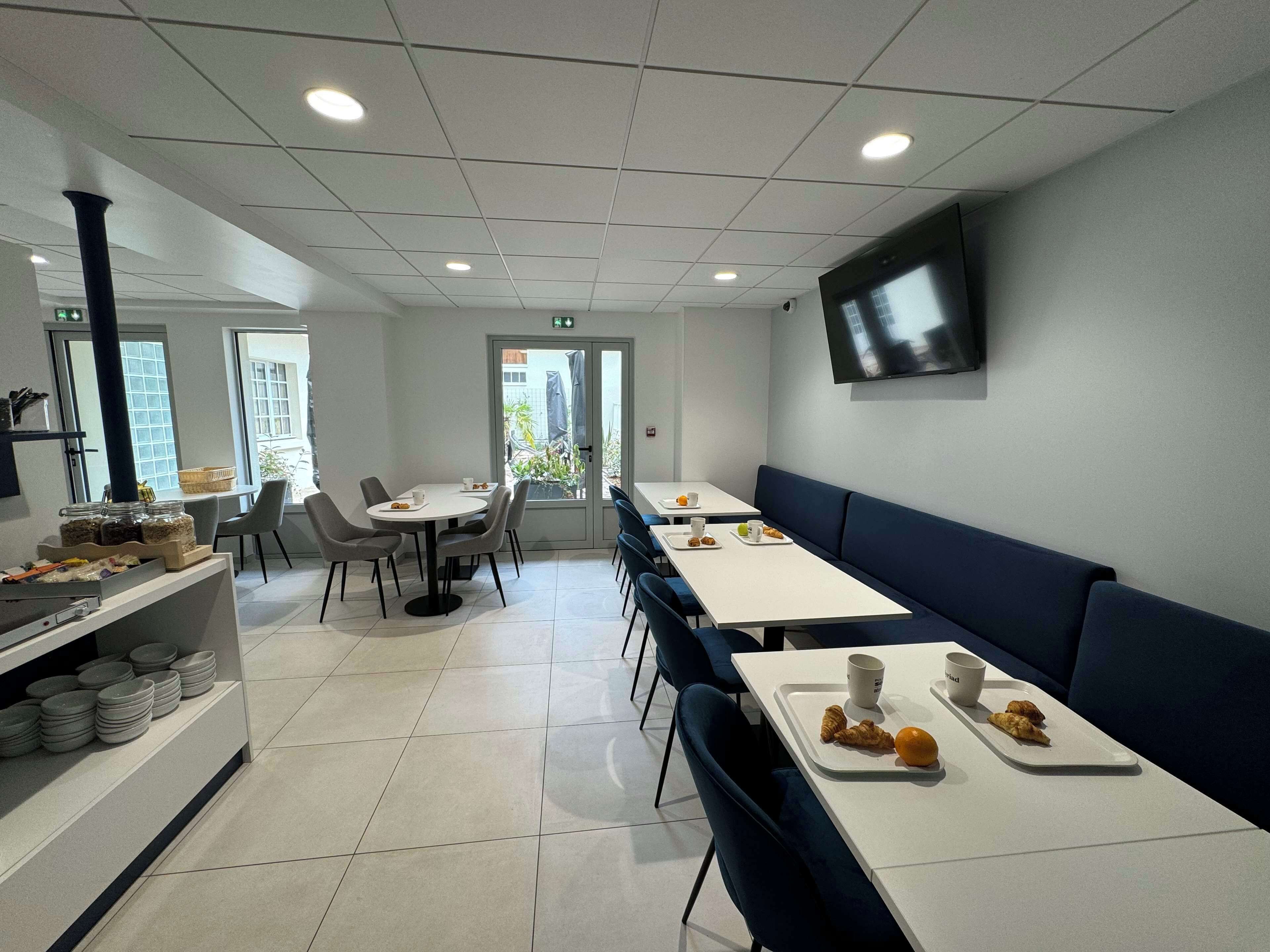 A modern dining area with white tables, blue chairs, and a long bench. There are plates with food and drinks on the tables.