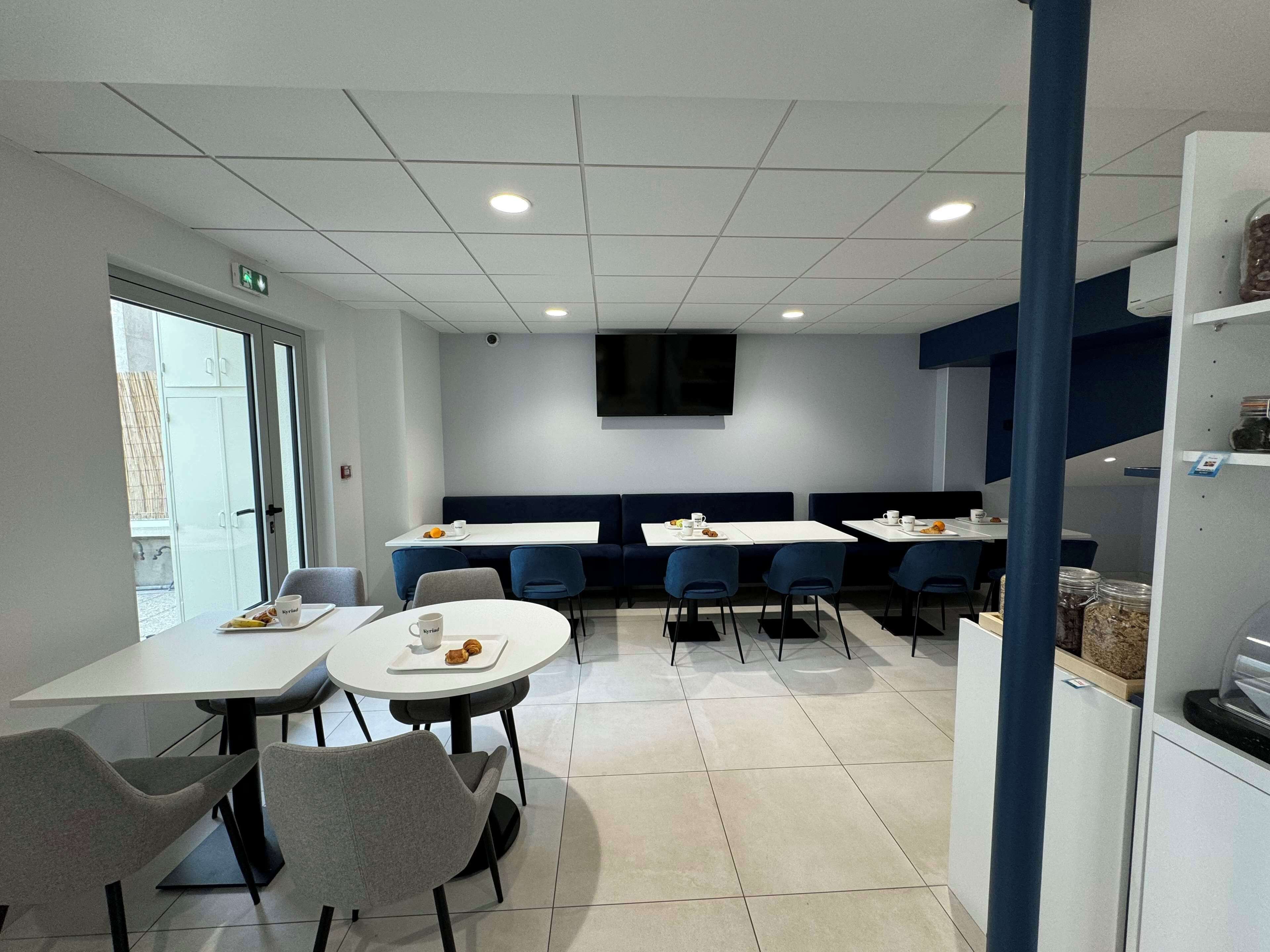 A modern dining area with white tables, blue chairs, and a flat-screen TV mounted on the wall.