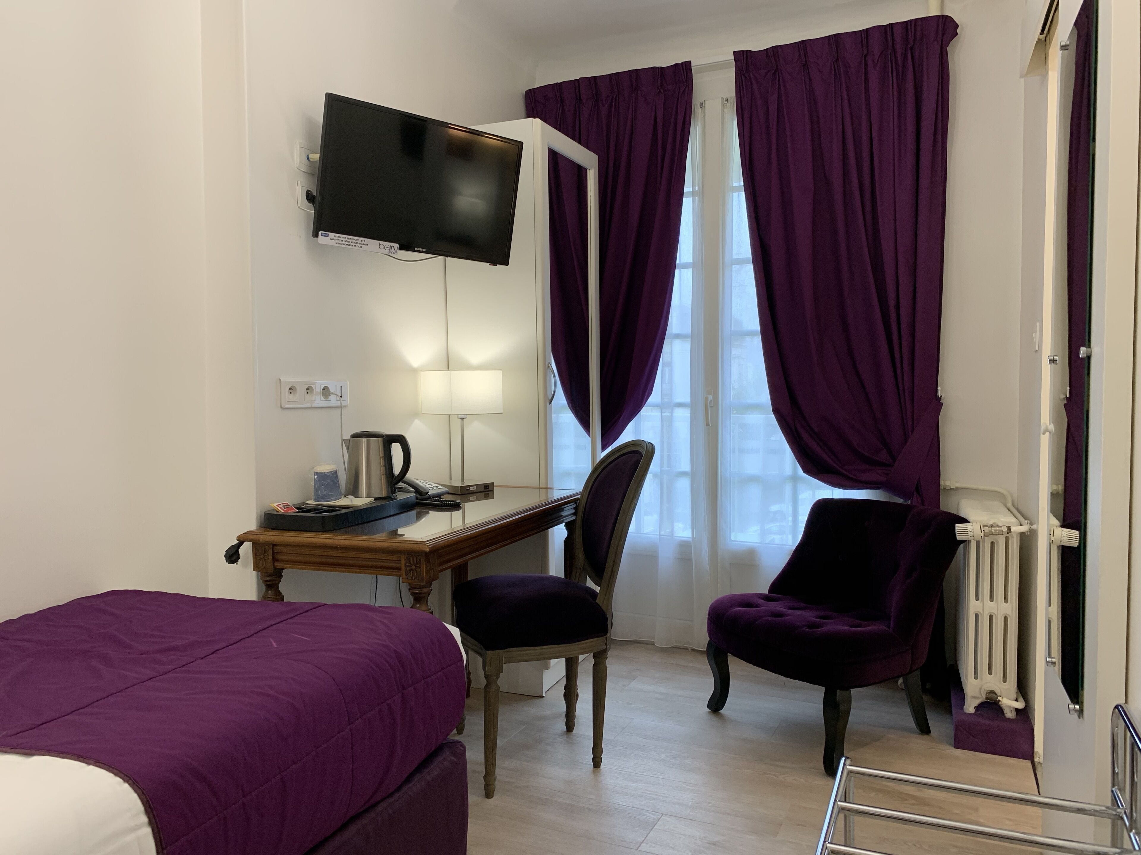 A hotel room with a bed, a desk with a computer, a chair, and a television on the wall.