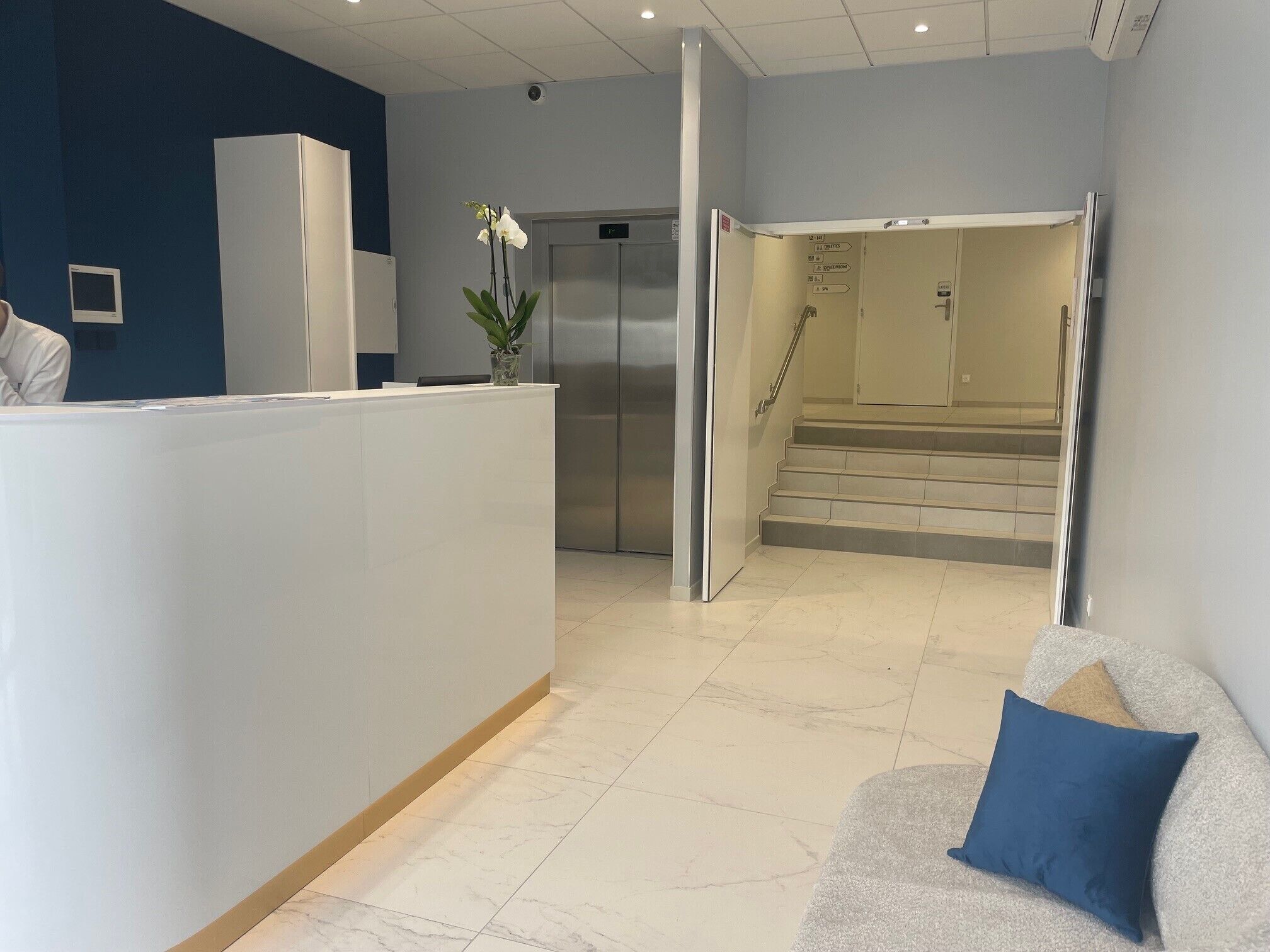 A reception area with a reception desk, a refrigerator, a staircase, and a waiting area with a couch and a blue pillow.