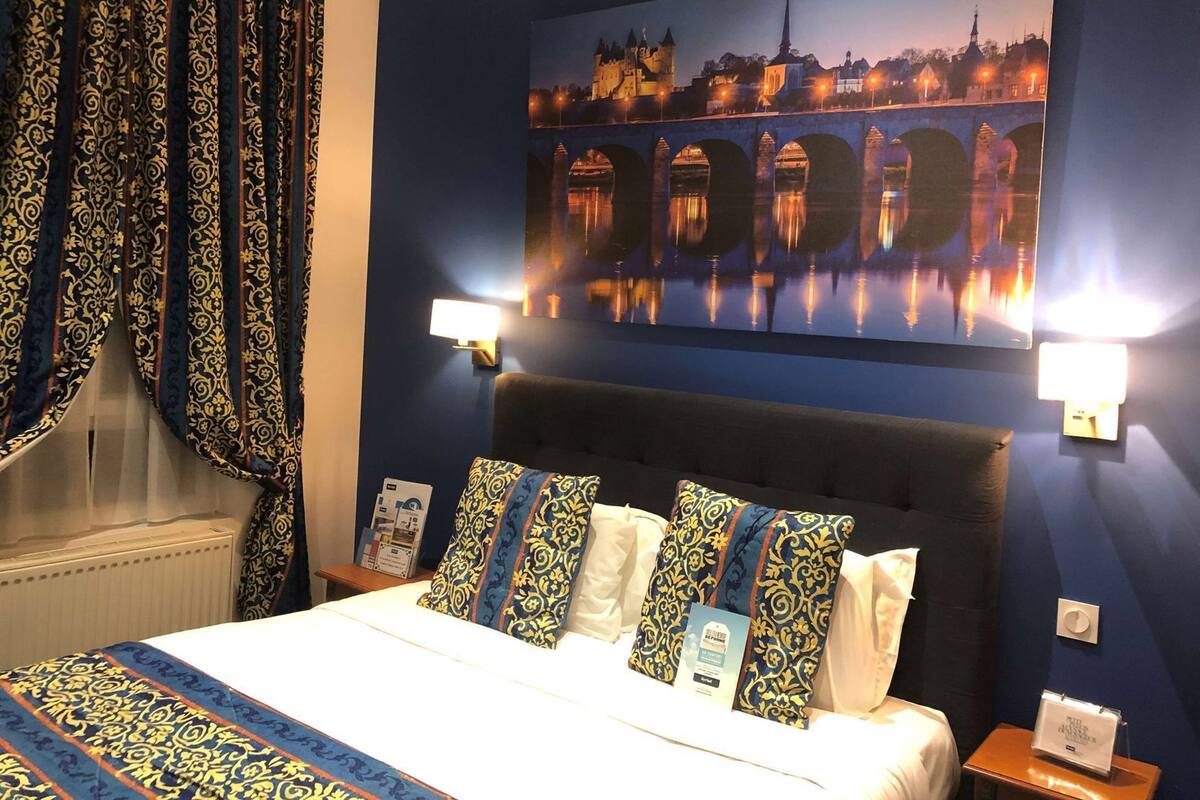 A hotel room with a bed, patterned pillows, a nightstand, a wall-mounted lamp, and a large cityscape painting.