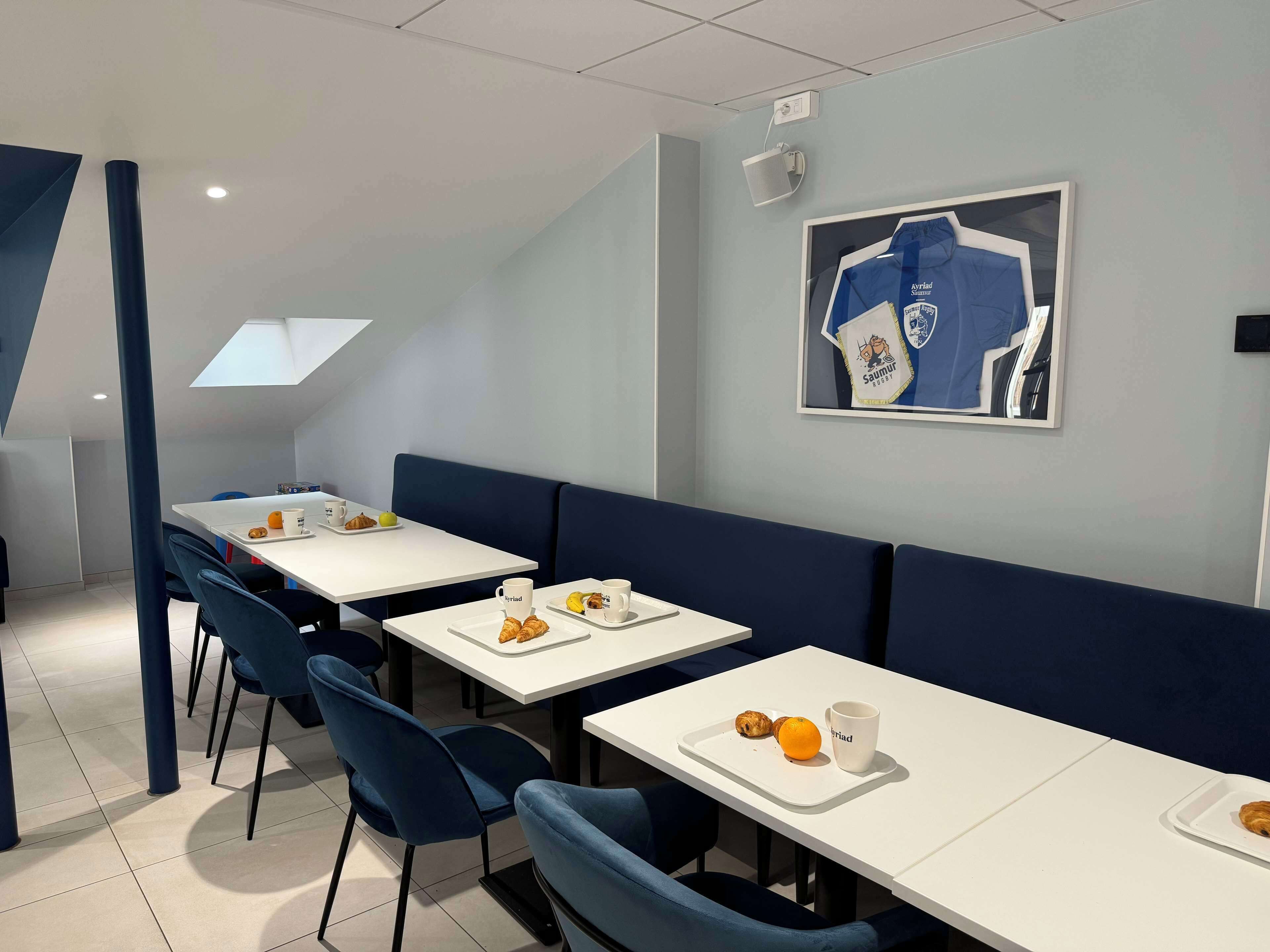 A modern dining area with white tables and blue chairs, featuring breakfast items like croissants and oranges.