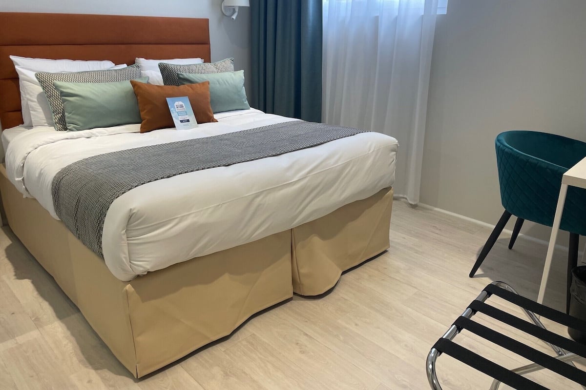 A neatly made bed with a headboard, pillows, and a blanket. There is a chair and a table to the right.
