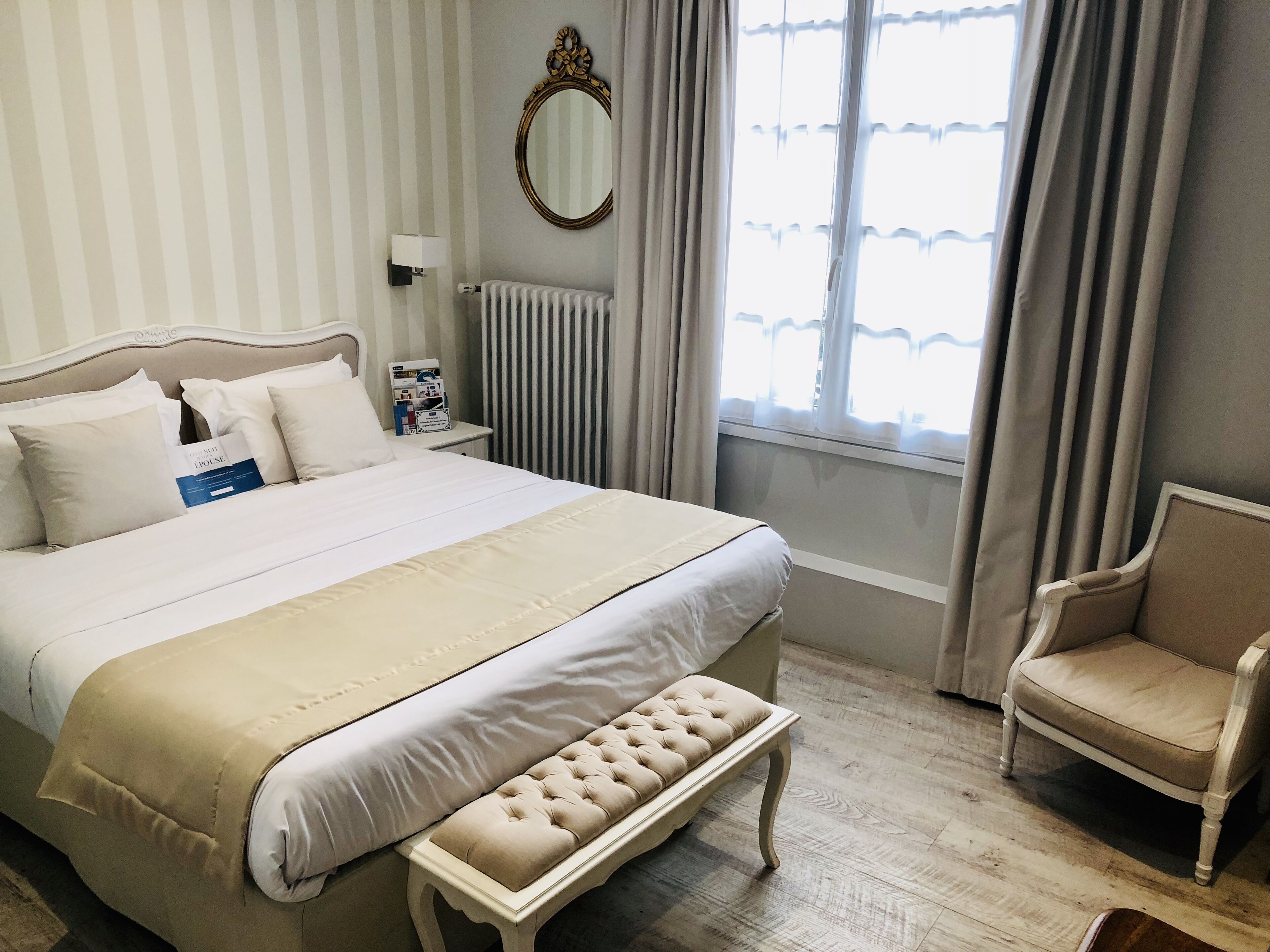 A neatly arranged bedroom with a large bed, a bedside table with a lamp, a chair, a window with curtains, and a radiator.