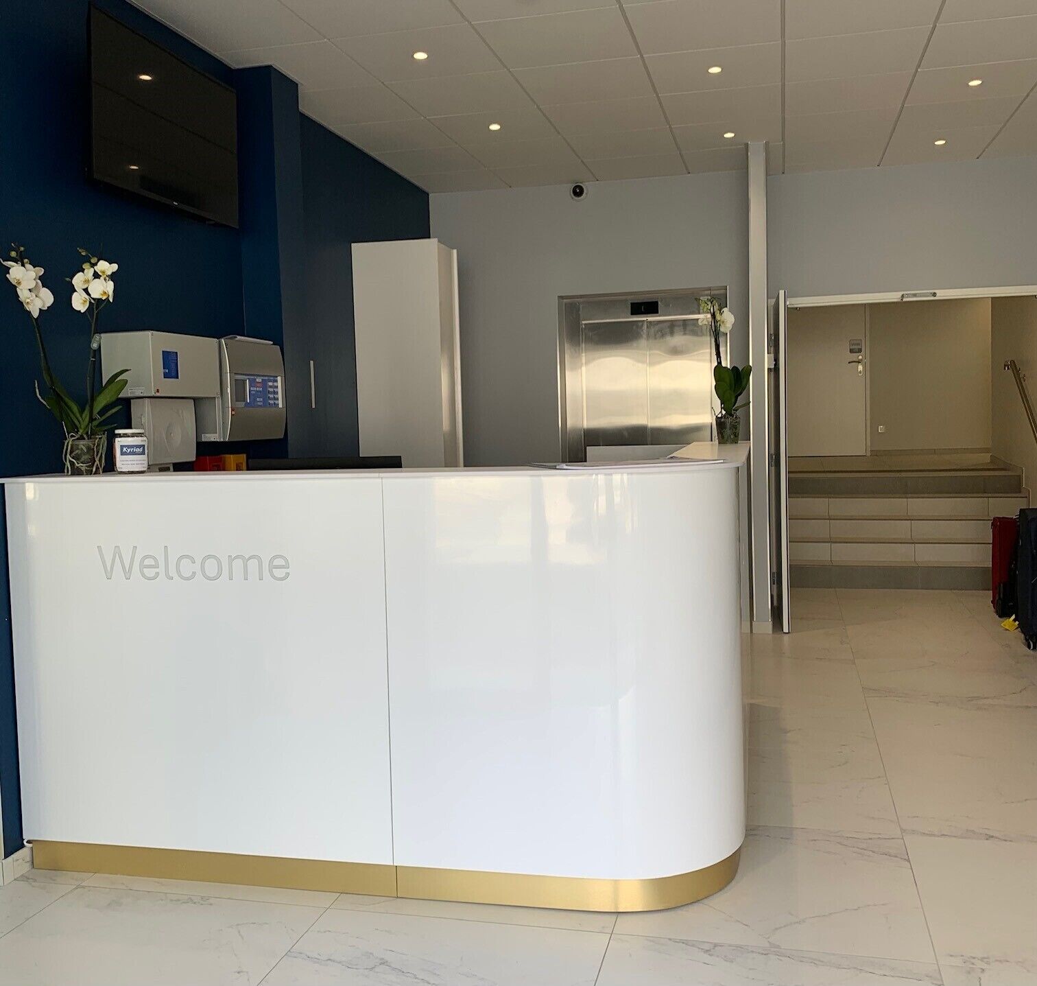 A reception desk with a 'Welcome' sign, a flat-screen TV, and a staircase leading to an upper floor.
