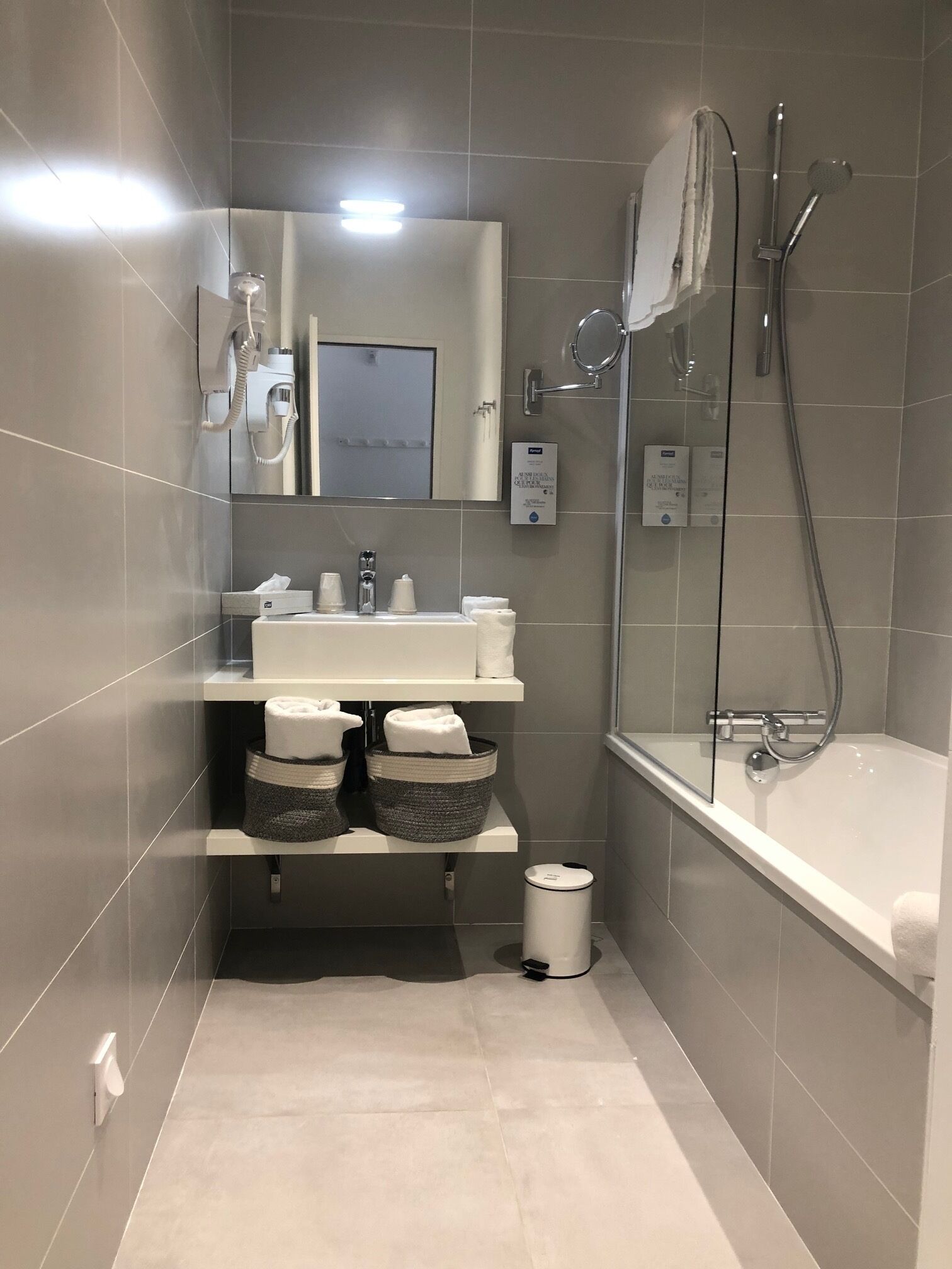 A modern bathroom with a white sink, mirror, and shower. There are two baskets under the sink and a trash bin next to it.
