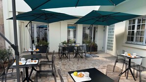 Un área de patio al aire libre con mesas y sillas, cubierta por grandes sombrillas y rodeada de plantas en macetas.