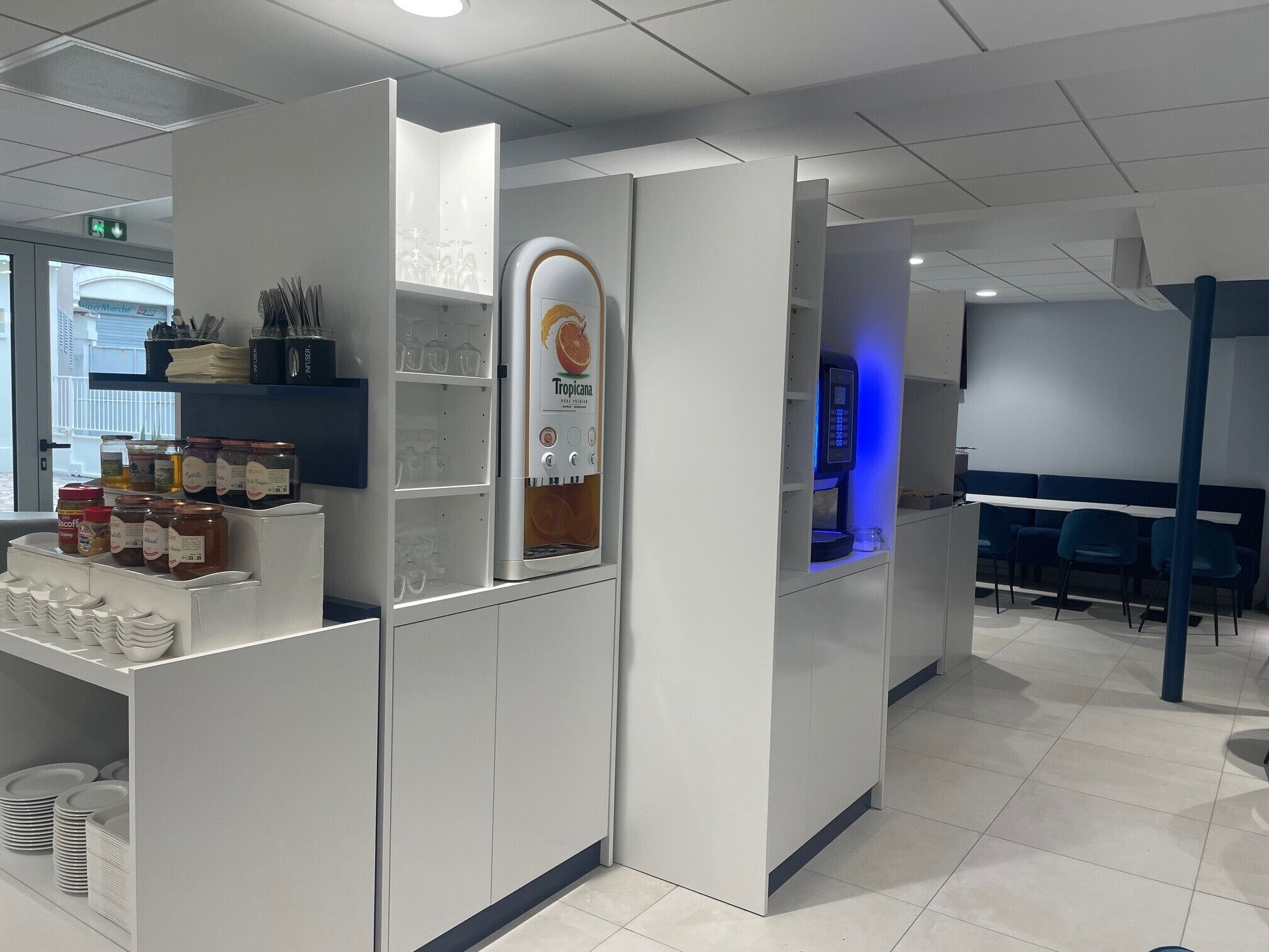 Una hall d'albergo moderna e luminosa, con un angolo bevande self-service, una zona relax con poltrone blu e vista sulla strada.