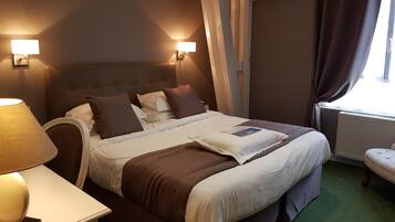 A hotel room with a bed, two bedside lamps, a nightstand, a window with curtains, and a radiator.