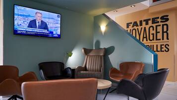 A modern lounge area with comfortable seating, a flat-screen TV displaying a news broadcast, and a wall with the text "FAITES VOYAGER PILES".