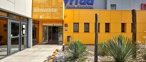 Entrada de un hotel con fachada amarilla y un letrero que dice 'Kyriad Hotel Restaurant'.
