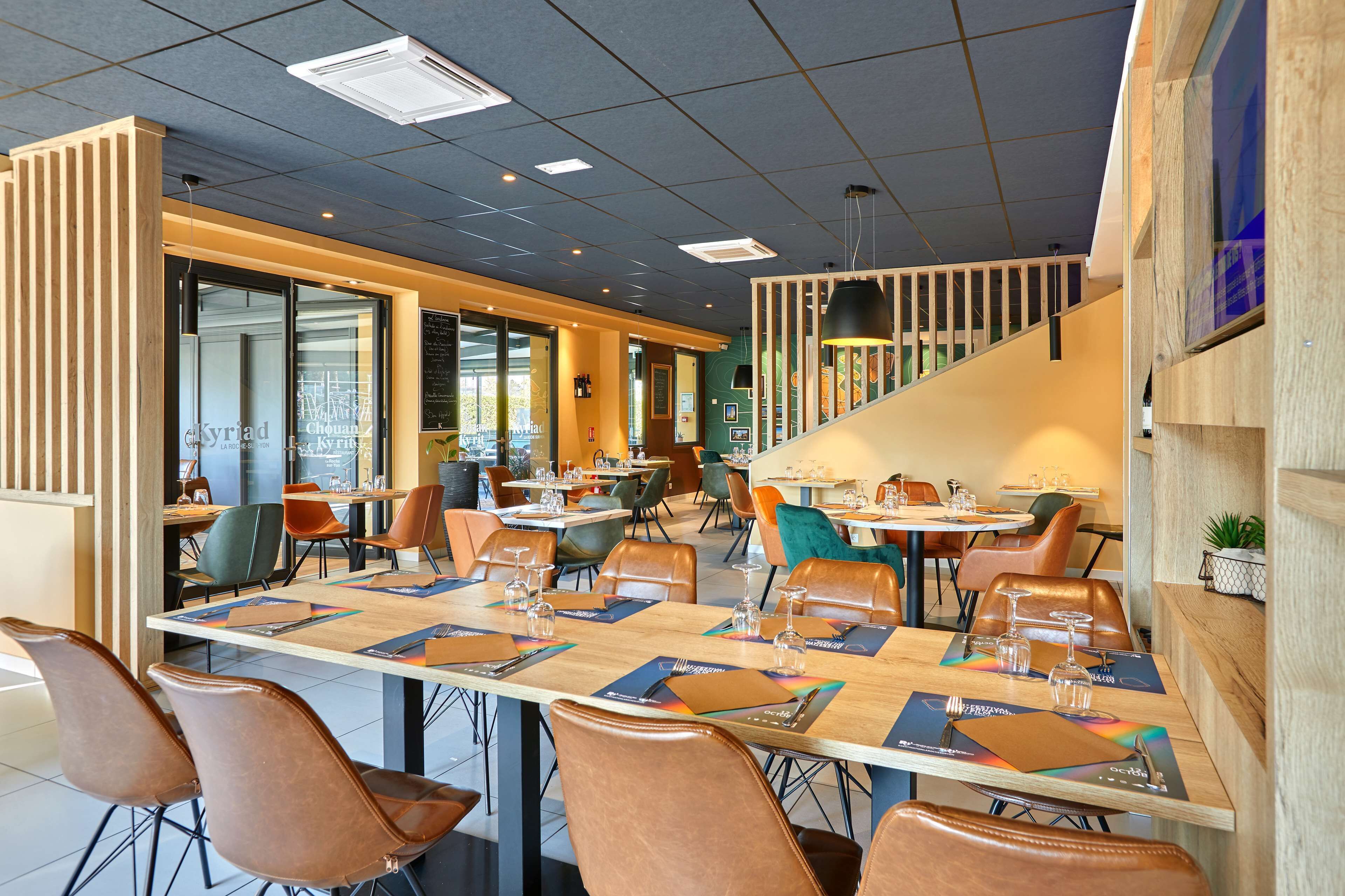 A modern restaurant with multiple dining tables, leather chairs, and a staircase leading to an upper level.