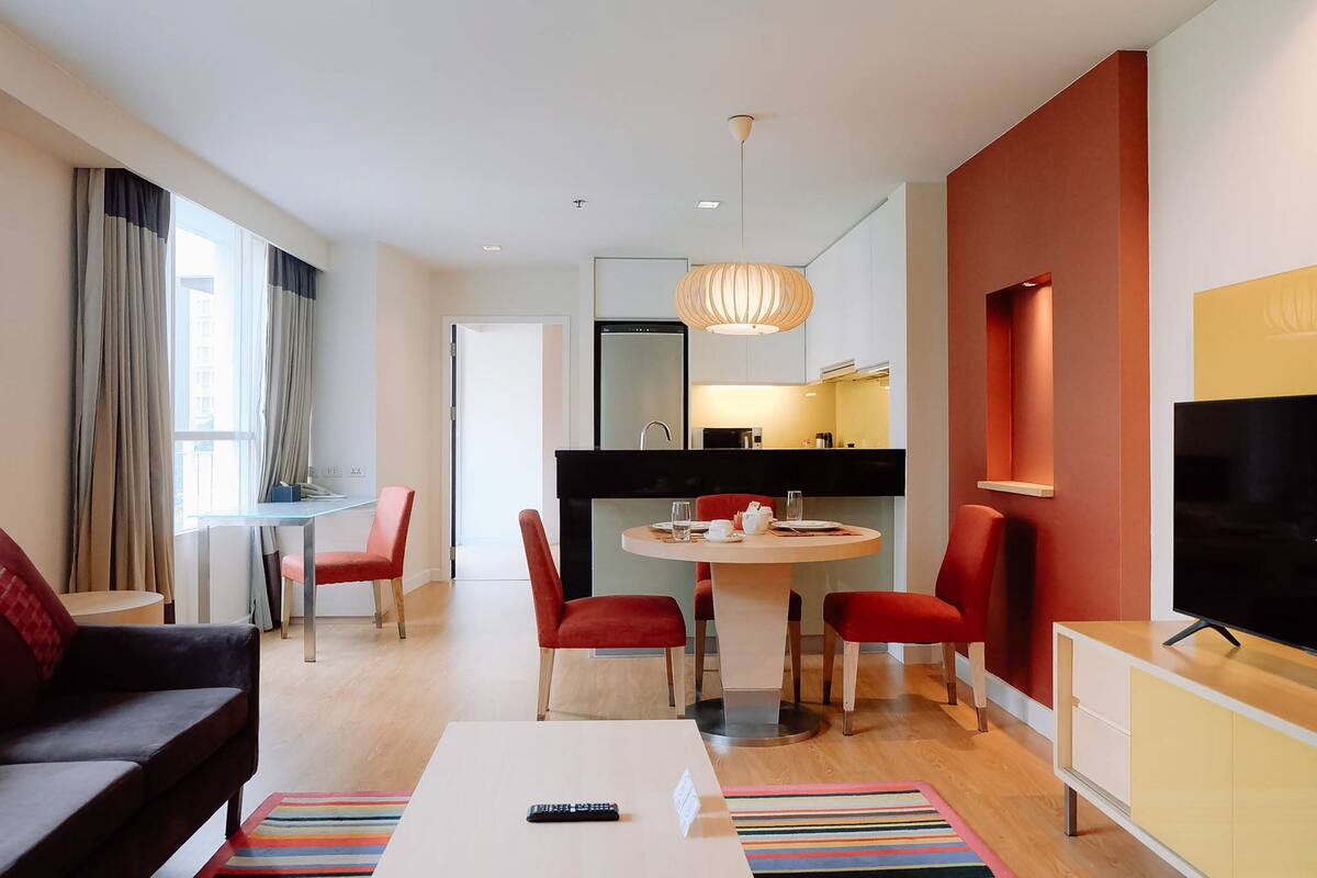 A modern hotel room with a dining area, a sofa, a TV, and a kitchen in the background.