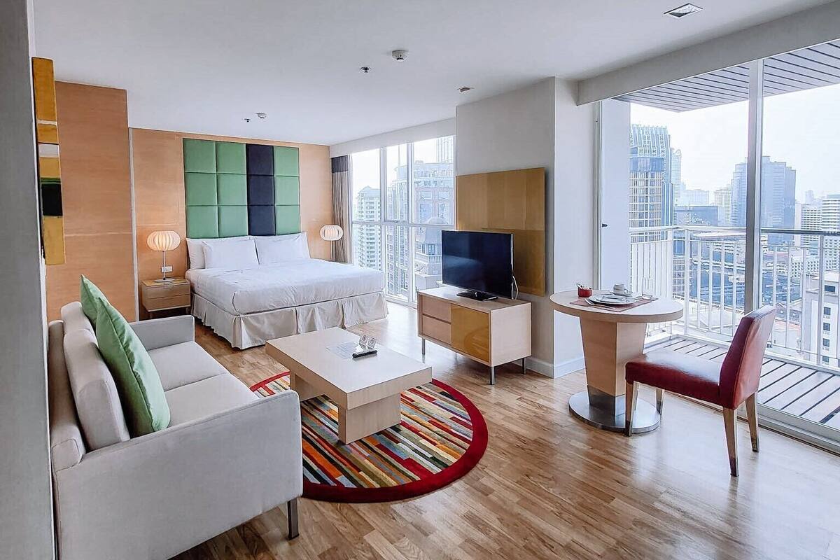 A modern hotel room with a bed, a desk, a chair, a TV, and large windows offering a city view.
