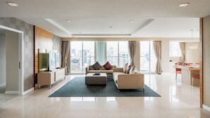 A spacious living area with a sofa set, a coffee table, and a TV on a stand. There is a large window with a city view, and a kitchen area is visible in the background. - Urbana Langsuan Hotel (Bangkok)