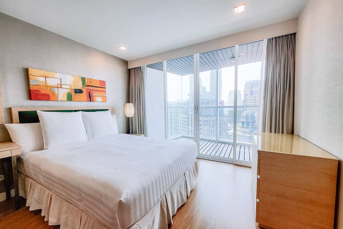 A modern hotel room with a large bed, a city view balcony, and a contemporary art piece on the wall.