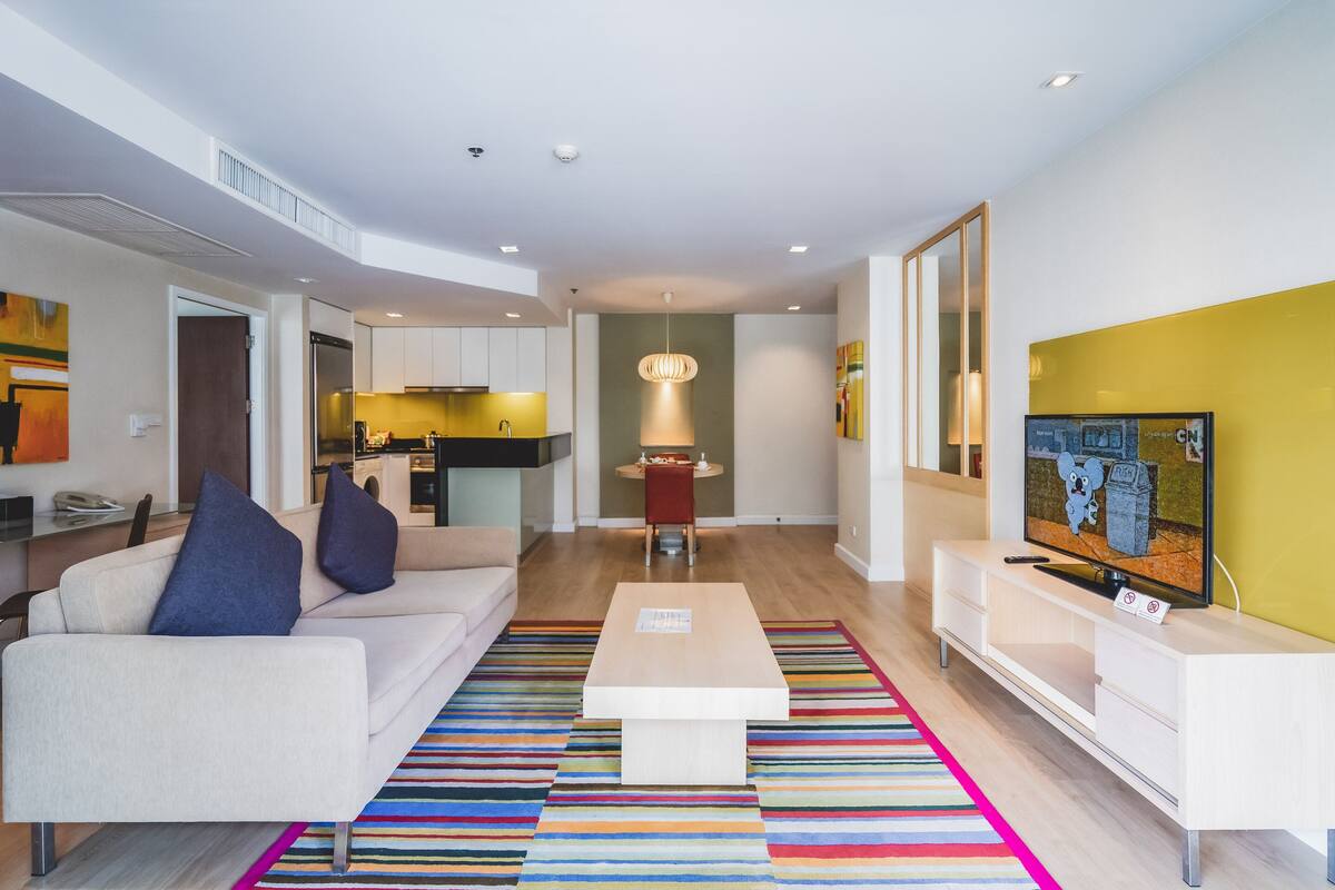 A modern living room with a sofa, coffee table, and TV. There is a kitchen in the background with a dining area.