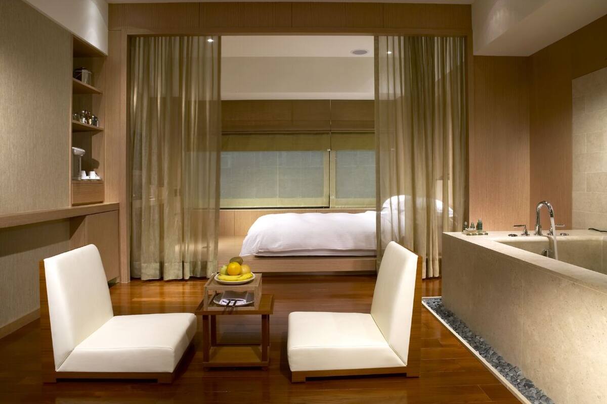 Hotelkamer met een bed, een zithoek met twee stoelen, een bijzettafeltje met een schaal fruit, een groot ligbad en een raam met gordijnen.