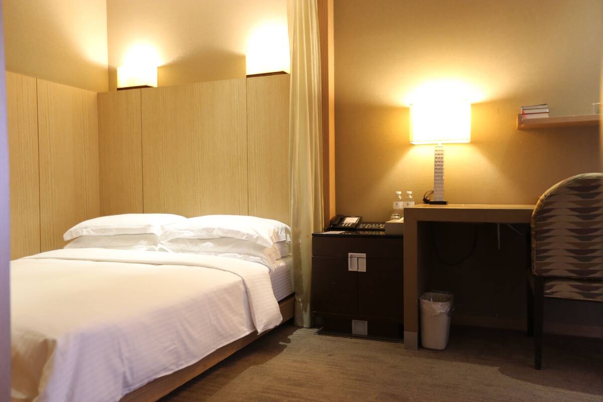 Een hotelkamer met een bed, een bureau met een lamp, een stoel en een prullenbak.