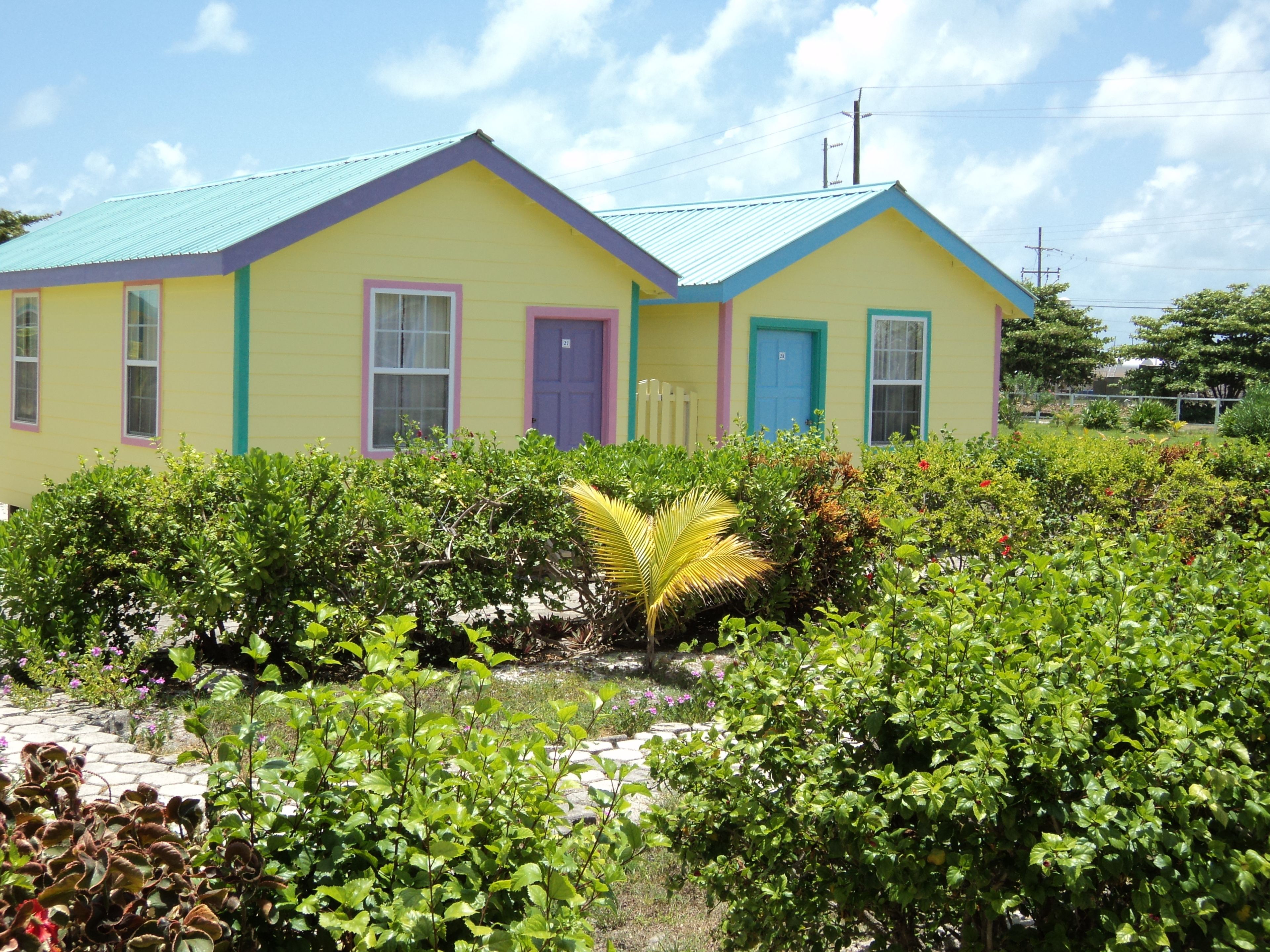A two-story building with a yellow facade and teal roof, surrounded by lush greenery.