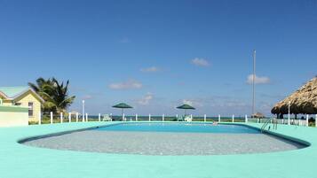 Una piscina con superficie de agua azul cristalina, rodeada por un suelo azul claro y barandillas blancas. Hay dos sombrillas verdes que dan sombra, y una estructura de techo de paja a la derecha.