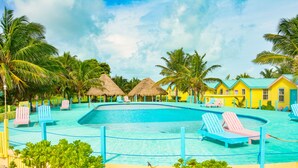 Zona de piscina con sillones de descanso, cabañas de techo de paja y palmeras.