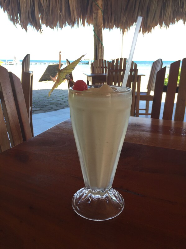 A tall glass of a creamy beverage with a cherry and a straw, placed on a wooden table at a beachside restaurant with wooden chairs and a thatched roof. - Royal Caribbean Resort (San Pedro)