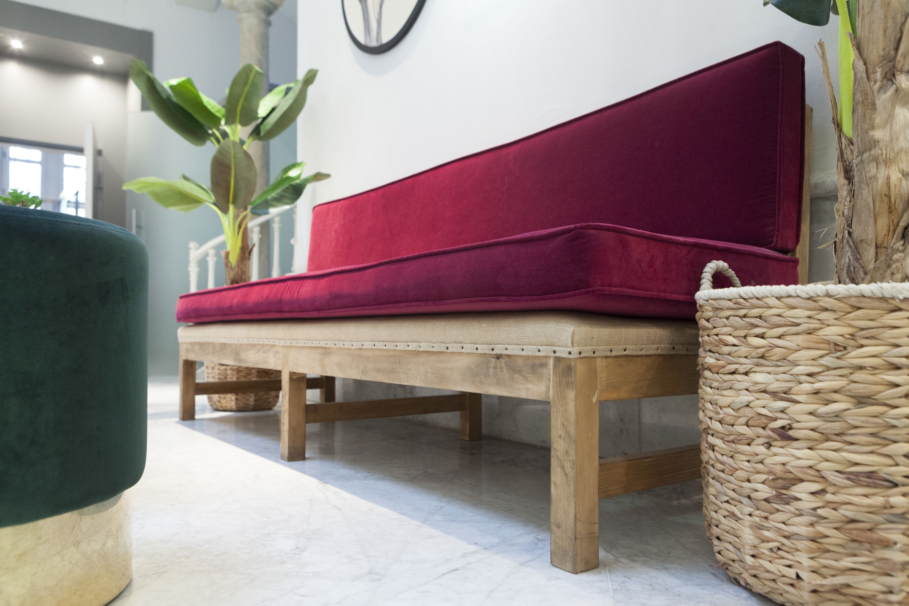 Un banco de madera con cojín rojo, una cesta tejida y una planta verde en una habitación moderna con pared blanca y un reloj.
