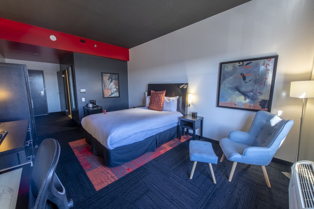Habitación de hotel con una cama grande, un escritorio, una silla, una lámpara de escritorio y obras de arte en las paredes.