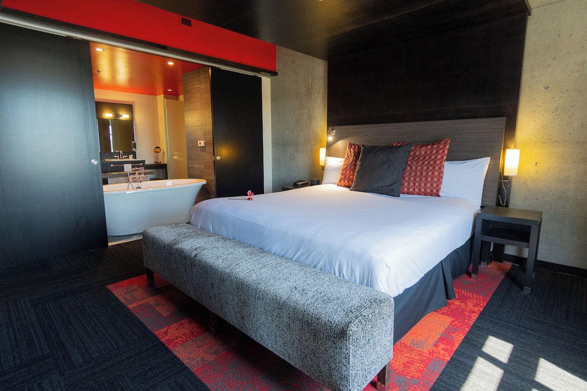 Habitación de hotel moderna con una cama grande, un banco, una mesita de noche pequeña, una mesita auxiliar, una lámpara de pared y un baño visible a través de una puerta abierta.