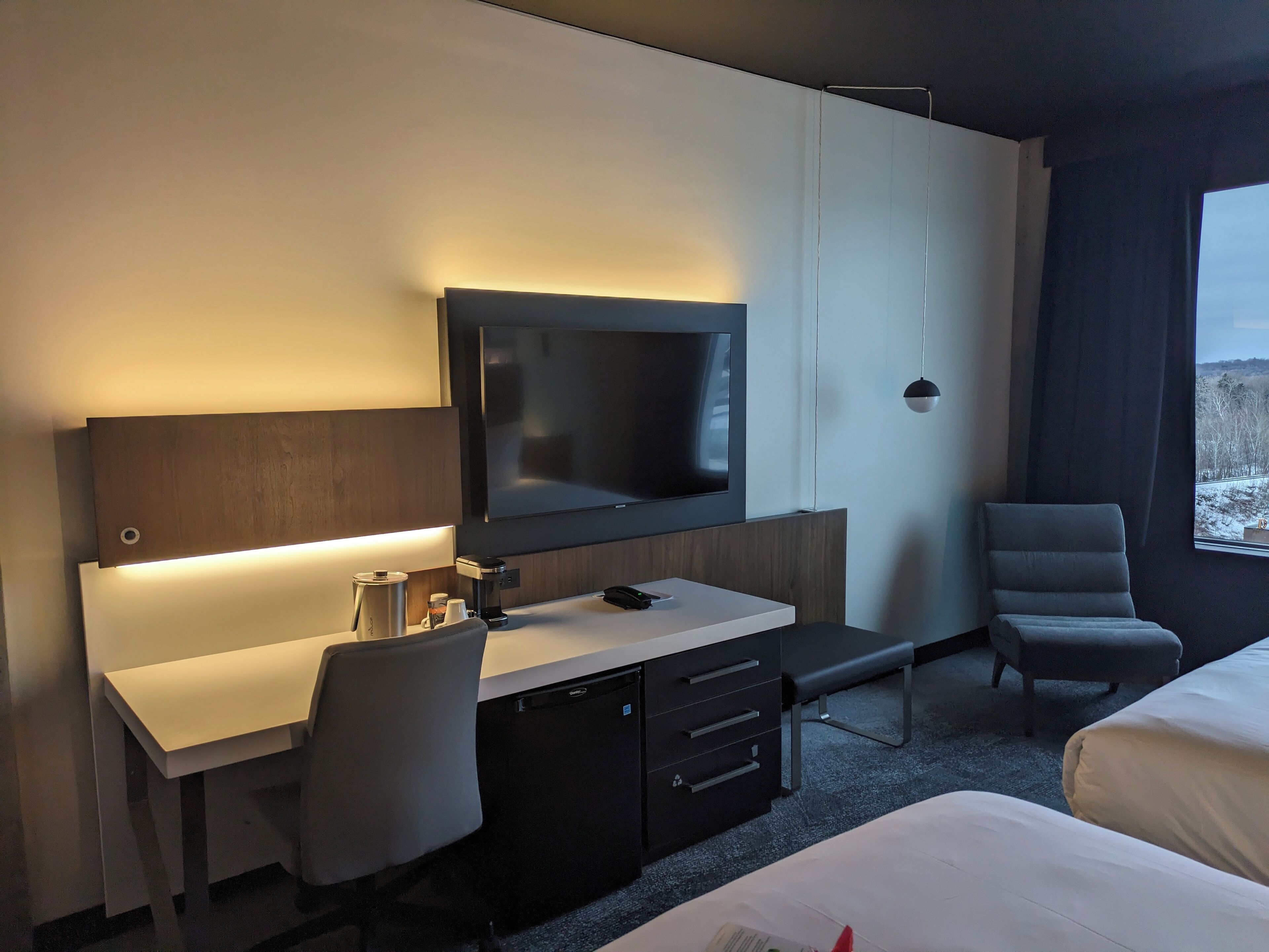 A modern hotel room with a flat-screen TV mounted on the wall, a desk with a chair, a bench, and a bed.