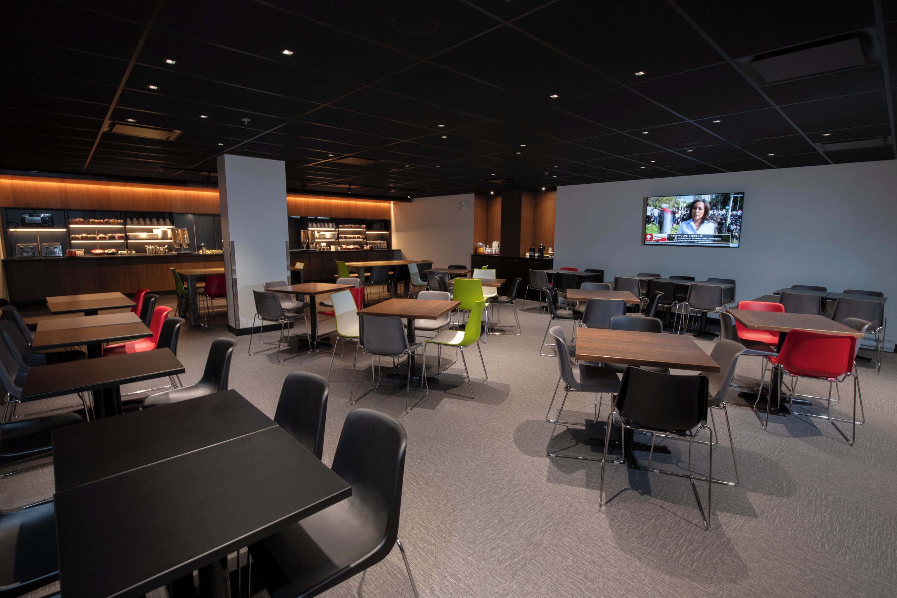 Een moderne congreszaal met tafels en stoelen, een bar en een groot scherm waarop een nieuwsuitzending draait.